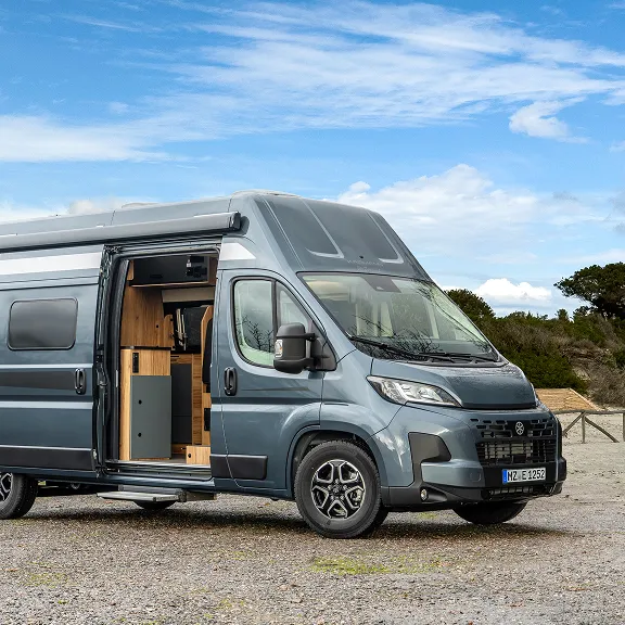 Grauer Campervan mit geöffneter Schiebetür steht am Ufer; Blick auf den Innenausbau und blauen Himmel.