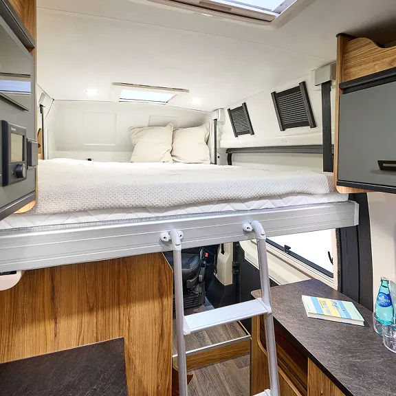 Innenansicht Campervan: festes Bett über dem Fahrerhaus mit Leiter, daneben Sitzgruppe und Stauraum.